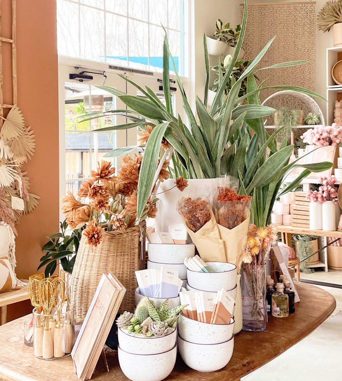 La boutique en ligne de Pigment présente une table remplie de plantes en pot et de vases, créant une atmosphère pleine de vie. Il y a plusieurs récipients sur la table, chacun contenant différentes plantes ou fleurs. La scène montre une collection diverse de plantes, certaines étant placées près de la fenêtre, ce qui contribue à créer un espace chaleureux et accueillant.
