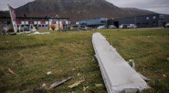 Betongbjelke ligger på grønn plen. I bakgrunnen ses en ramponert bygning foran et fjell.