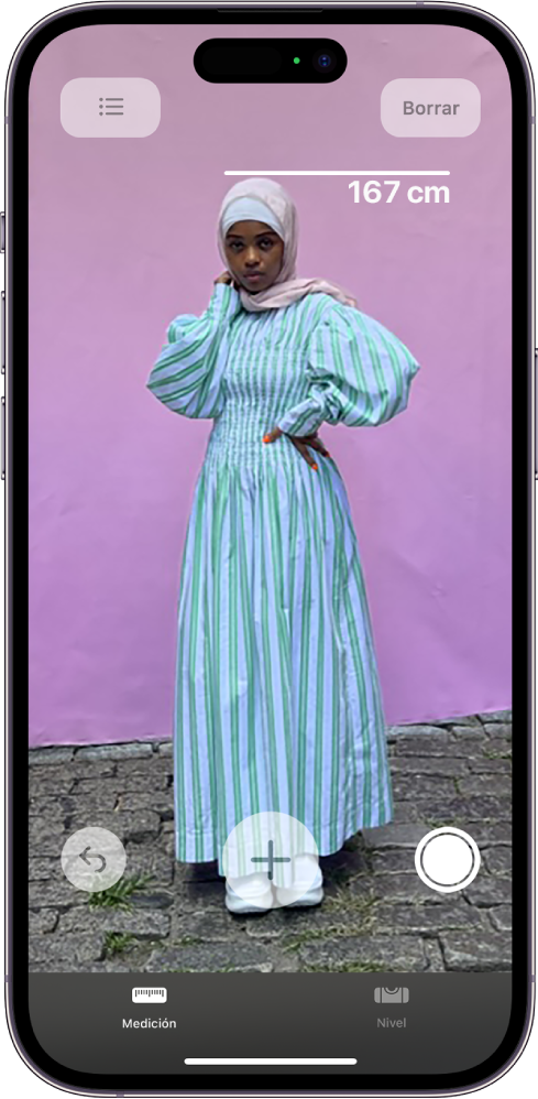 Se mide la altura de una persona y se muestra la medición sobre su cabeza en la app Medición. El botón Tomar foto está activo cerca de la esquina inferior derecha para tomar una foto de la medición. El indicador verde Cámara en uso aparece en la parte superior.