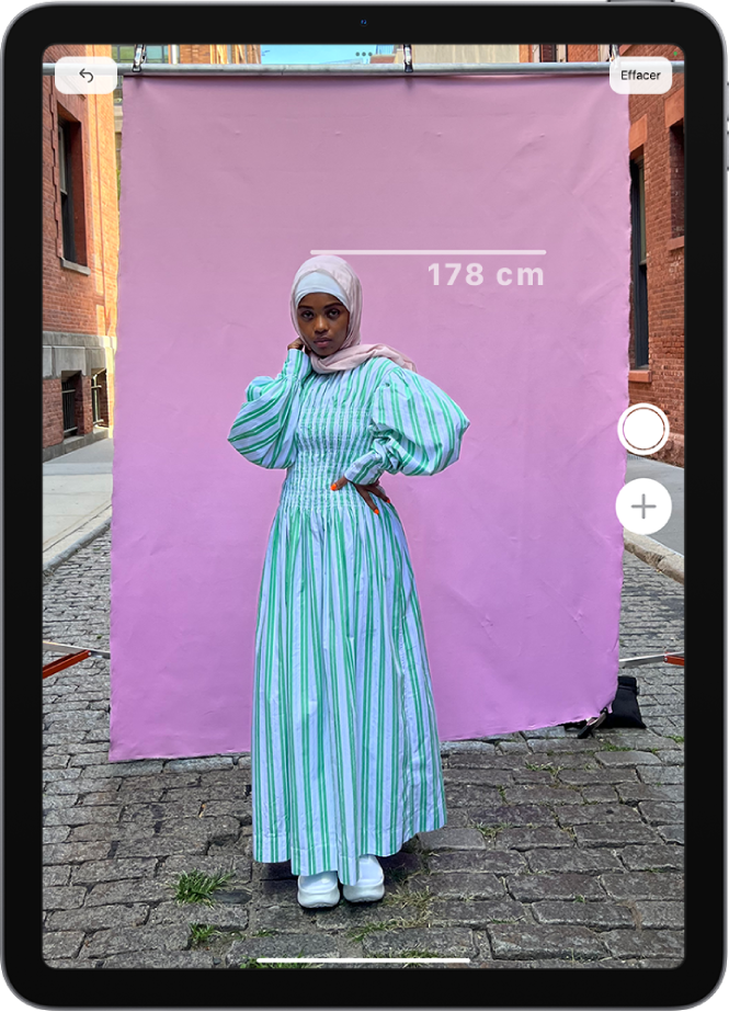 La taille d’une personne est mesurée dans l’app Mesure, la mesure apparaissant au-dessus de la tête de la personne. Le bouton « Prendre une photo » est actif en bas à droite pour prendre une photo de la mesure. Le voyant vert d’appareil photo en cours d’utilisation apparaît en haut.