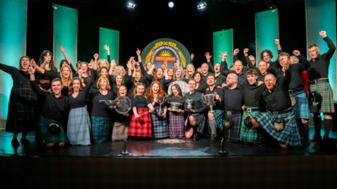 Còisir sa bheil boireannaich agus fireannaich air an èideadh ann am fèilidhean tartain a' dèanamh gà irdeachais le an gà irdeanan suas. Tha iad seachdnar air an glùinean agus cupanan agus cuachan a bhuannaich iad aca