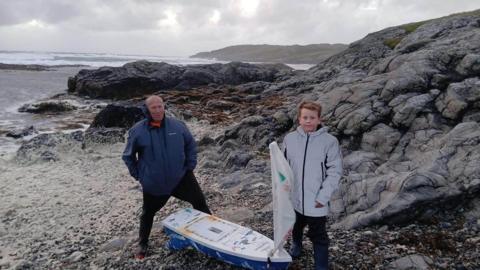 Balach beag le seacaid liath agus fireannach le seacaid ghorm nan seasamh ri taobh bà ta beag le seòl air cladach creagach air là glas.