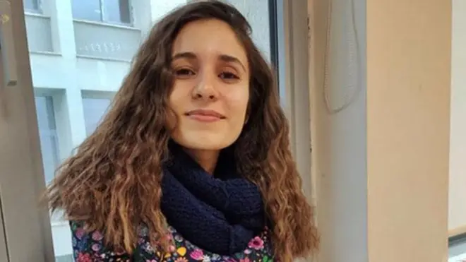 Gülistan Doku uzun kıvırcık saçları ve çiçekli elbisesi ve koyu renk atkısı ile kameraya bakarak gülümsüyor