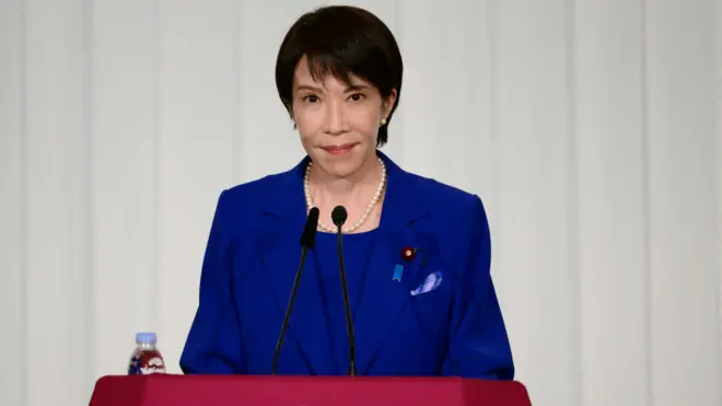 Санаэ Таикити с короткой стрижкой, в ярко-синем костюме и с жемчужным орежельем стоит перед микрофонами на пресс-конференции.