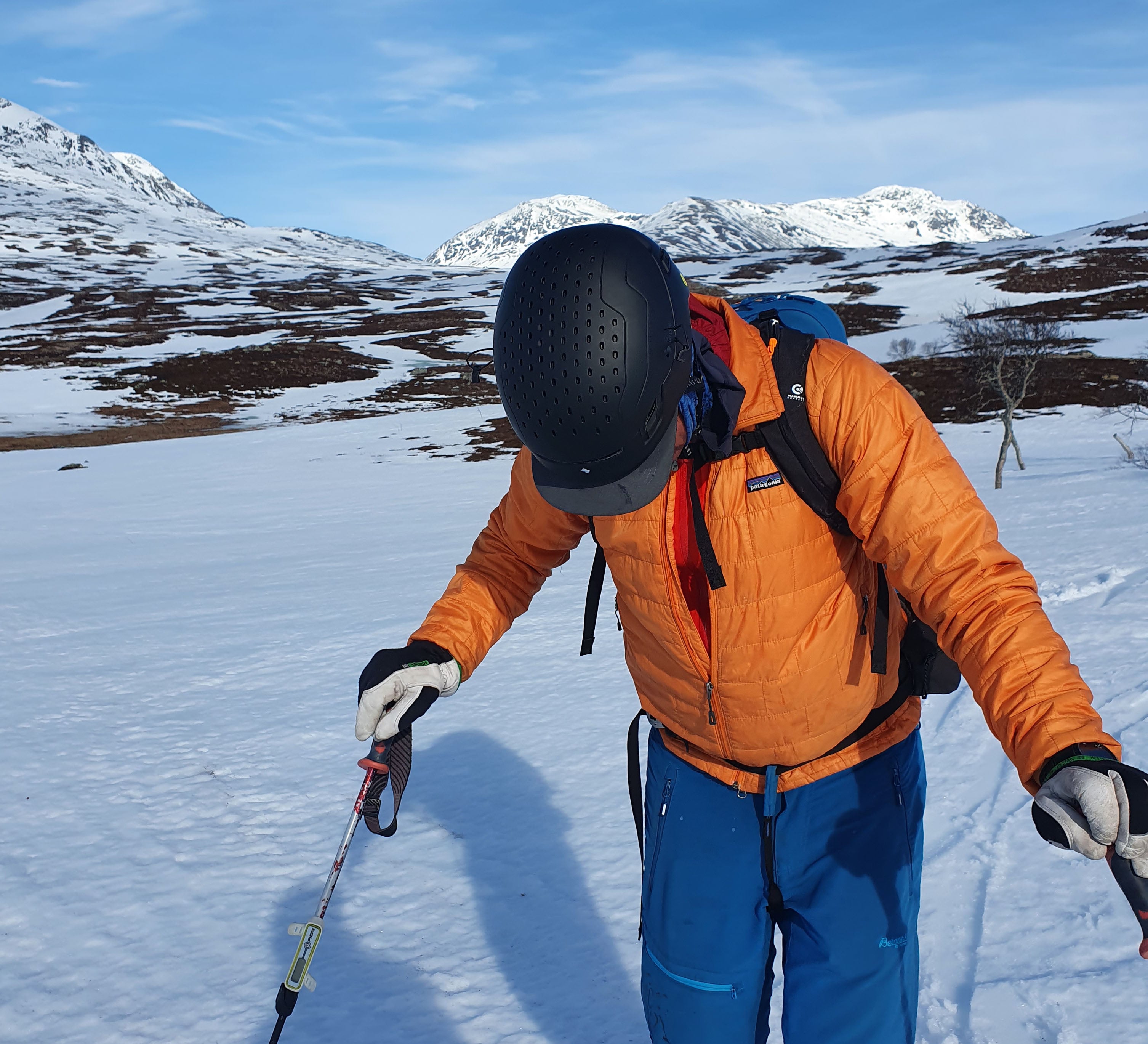 Skidåkare med orange jacka blåa byxor står stilla och tittar ner i snön. Man ser inte ansiktet utan bara den svarta hjälmen.