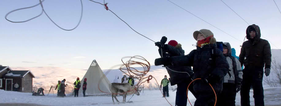 Foto tatt utendørs med snø på bakken og solnedgang. Noen unge gutter kaster lassoer opp i luften foran seg. I bakgrunnen er et reinsdyr, noen voksne med refleksvester, en lavvo og et lite rødt hus.