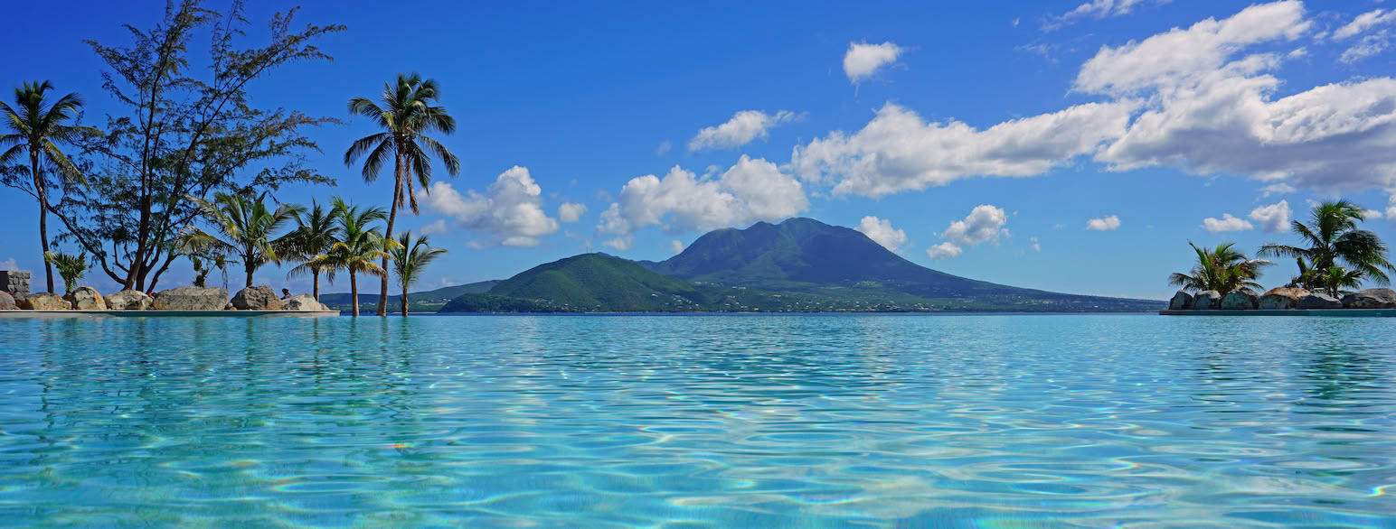 I forgrunnen palmer og blått hav, i bakgrunnen en vulkan på en annen øy.