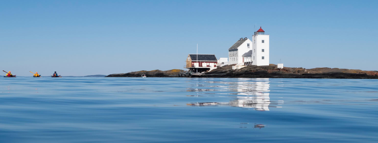 et fyr med en hytte ved siden av ligger på en holme, badet i sol. Inn fra venstre kommer tre kajakker padlende. 