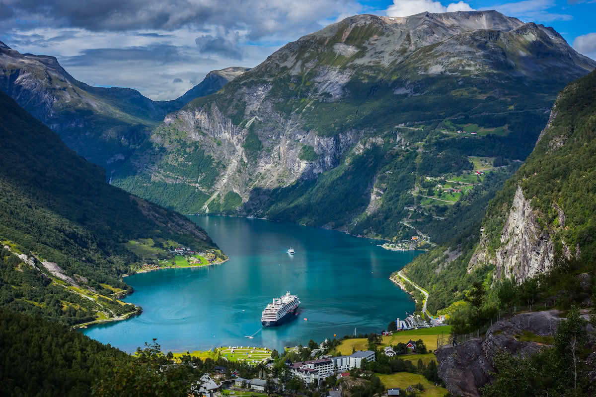 Et cruiseskip ligger på vannet. Nærmest kamera er et tettsted med et hotell og noen andre bygninger. Høye fjell ligger rundt fjorden, og på fjellet midt-i-mot tettstedet er en vei med mange svinger opp til noen hus i fjellsiden.