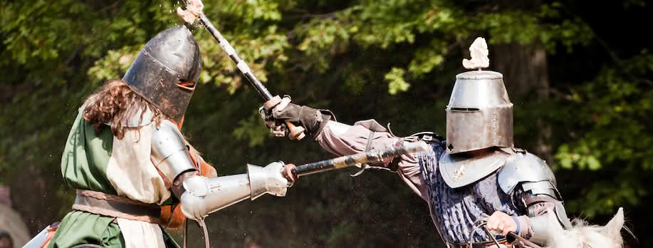 Bilde fra en moderne ridderturnering. To riddere i rustning som kjemper. Rustningen er først og fremst en hodebeskyttelse. De slåss mot hverandre med stokker. De sitter på hver sin hest. i bakgrunnen er det tilskuere.