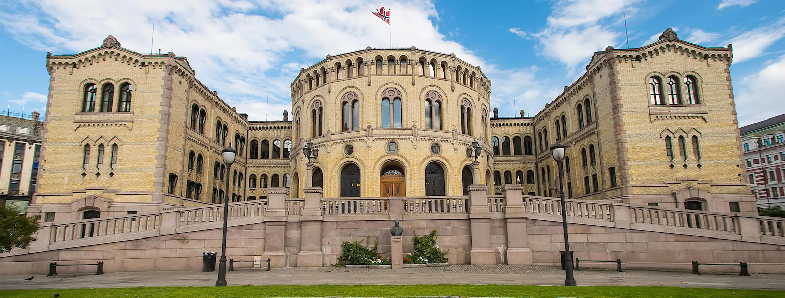 vaier et norsk flagg. Foran bygningen kan man gå opp en liten vei til inngangen. Veien er bygd opp med en mur, og har rekkverk i stein. Foran bygningen er en plen.