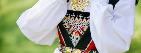 Nærbilde av ei kvinne med Fanabunad med perlebroderier foran på vesten og perlebrodert belte. Ho held armane opp, har kvit langerma bunadskjorte og ei stor gullfarga sølje over vesten. Foto