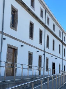 A long white building with wooden windows and doors, stone borders, and a sloped accessible pathway under a clear blue sky.
