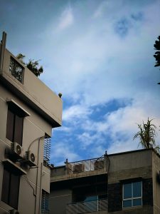A bright blue sky with soft white clouds rises above a cluster of modern concrete buildings. The left building has balconies, railings, and two wall-mounted air-conditioning units. Rooftops feature metal railings and several plants, including tall palms. Darker stone cladding surrounds a window on the right.