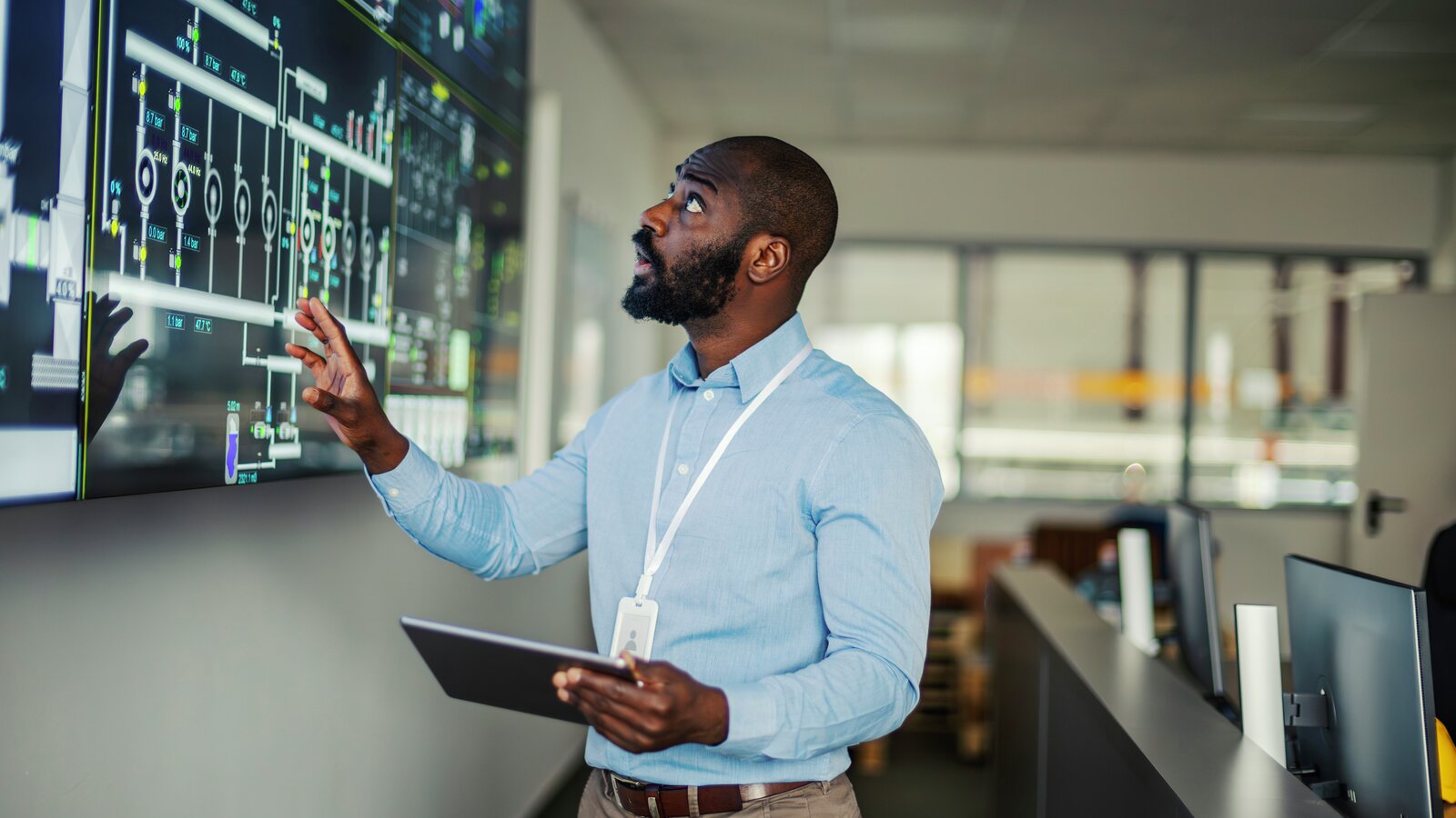 Ingeniør som holder et digitalt nettbrett og observerer komplekse data på en stor skjerm, og administrerer industriell drift i et moderne kontrollrom