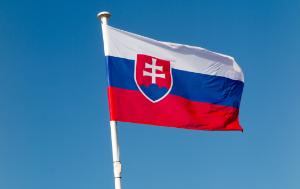 A flag with three horizontal stripes—white on top, blue in the middle, and red at the bottom—is waving against a clear blue sky. On the left side of the flag, there is a red shield with a white double cross standing on three blue hills. This is the national flag of Slovakia.