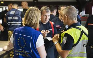 A group of emergency response professionals stand together indoors during what appears to be a coordination meeting or operational briefing. In the foreground, a person wearing a blue vest marked “European Union Civil Protection” speaks with two responders in uniform.