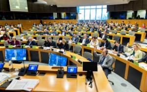 A large modern conference room at the European Parliament is filled with dozens of people seated in curved rows of desks. Many participants are voting by raising one hand, during a formal meeting. Most are wearing business attire and using microphones, laptops, or headsets. Large screens at the front display a man speaking remotely. Bright windows at the back let in daylight, giving the room a professional institutional atmosphere.