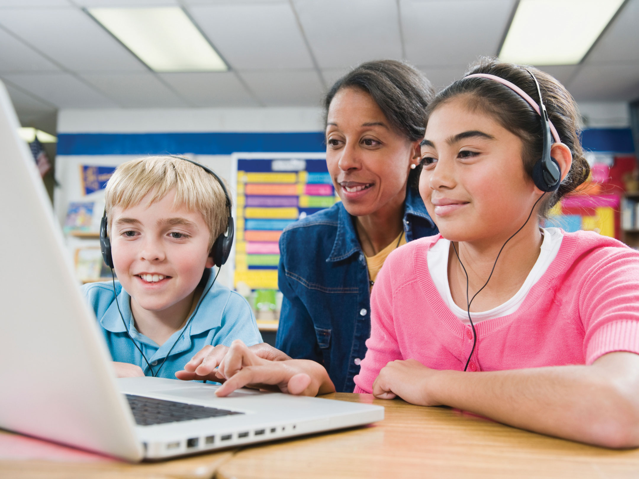 Two students at school wearing headphones using text-to-speech technology while doing their homework on a laptop. The teacher sits behind them watching how they work.