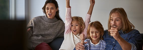 Et par med to små barn ser på TV og heier engasjert, det ene barnet strekker armene i været