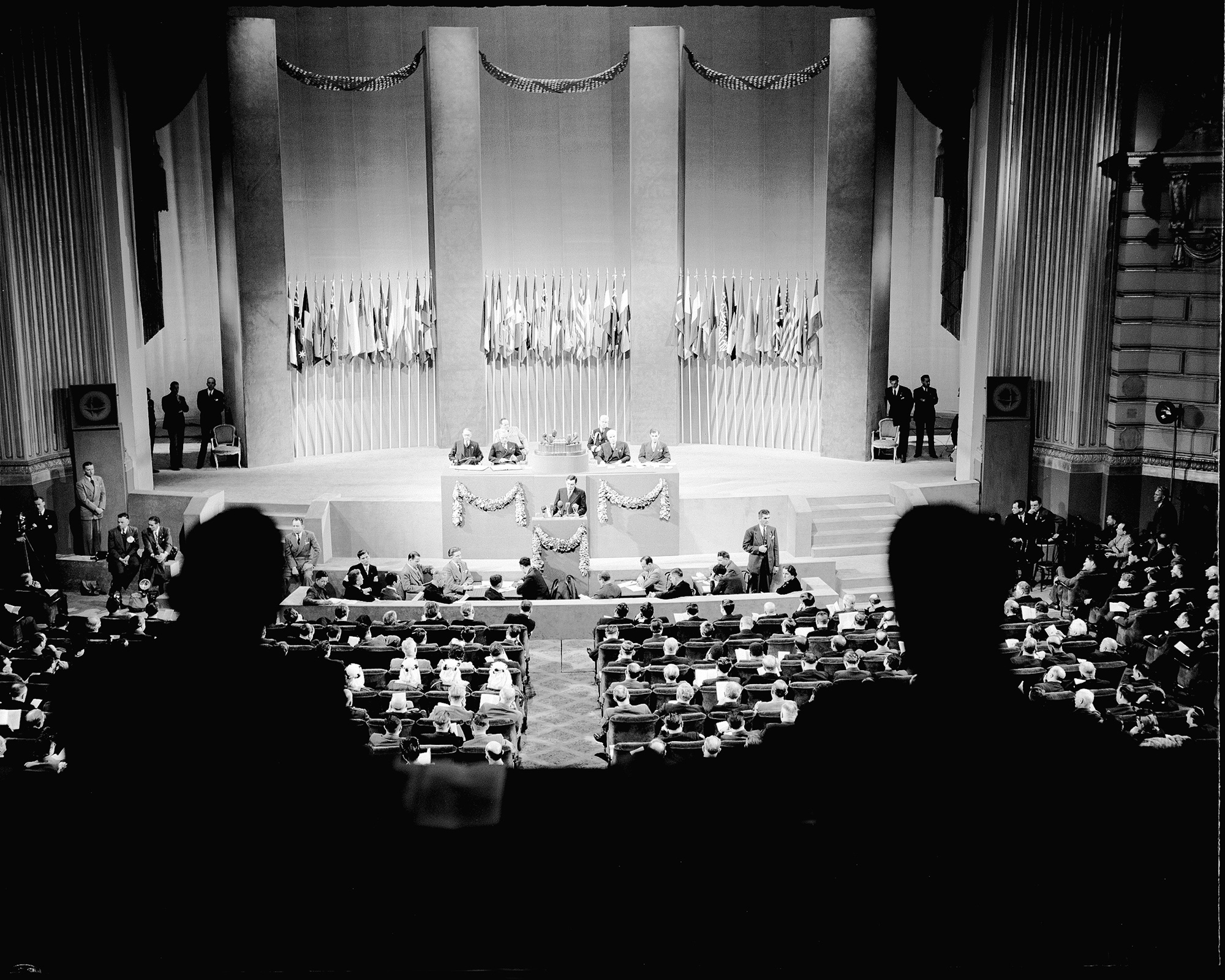 bird's eye view of many in attendance at a grand hall with tall columns and several flags
